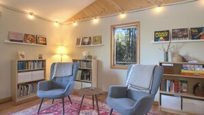 A cozy living room with two armchairs, a small round table, and a bookshelf filled with books. A window with a view of trees is visible, and there are string lights on the ceiling. - Boreal Retreat  |  Cabin in the Pines | Port Wing (Port Wing)