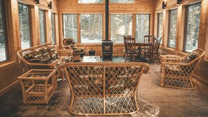 A log cabin interior with wicker furniture, a central fireplace, and large windows offering a view of the forest. - Boreal Retreat  |  Cabin in the Pines | Port Wing (Port Wing)