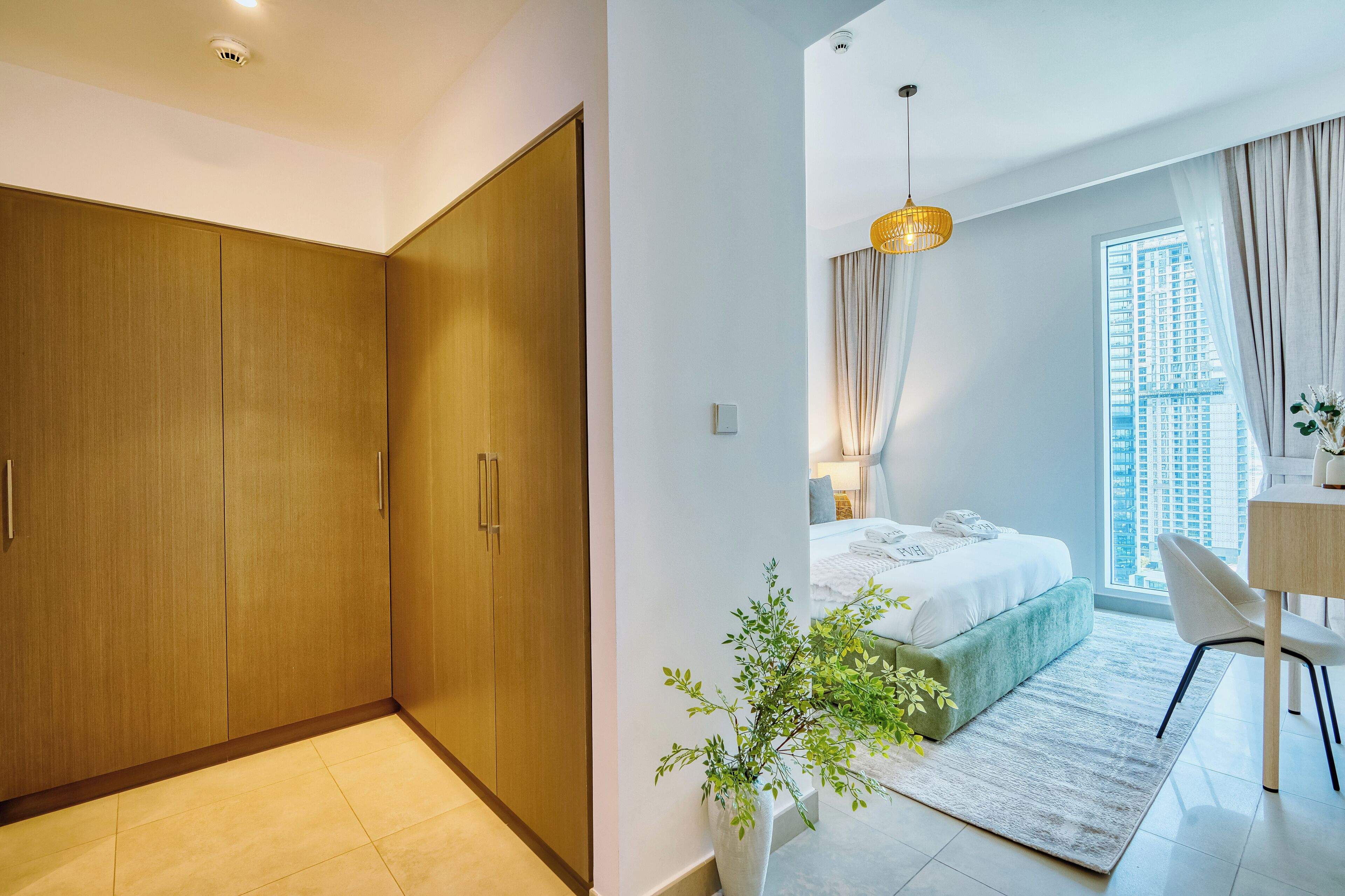 A modern bedroom with a large wardrobe, a bed with a green upholstered headboard, a small desk, and a view of a cityscape through the window.
