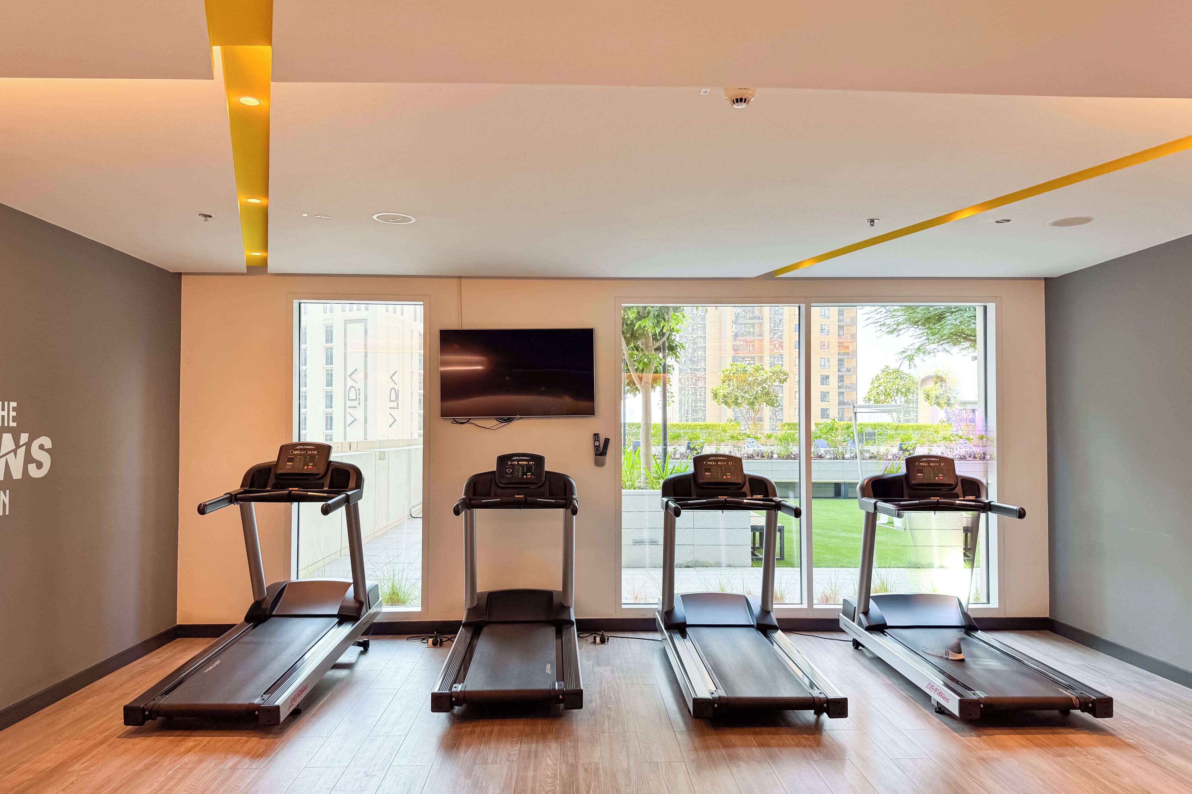 Ein Fitnessraum mit vier Laufbändern, großen Fenstern und einem Flachbildfernseher an der Wand.