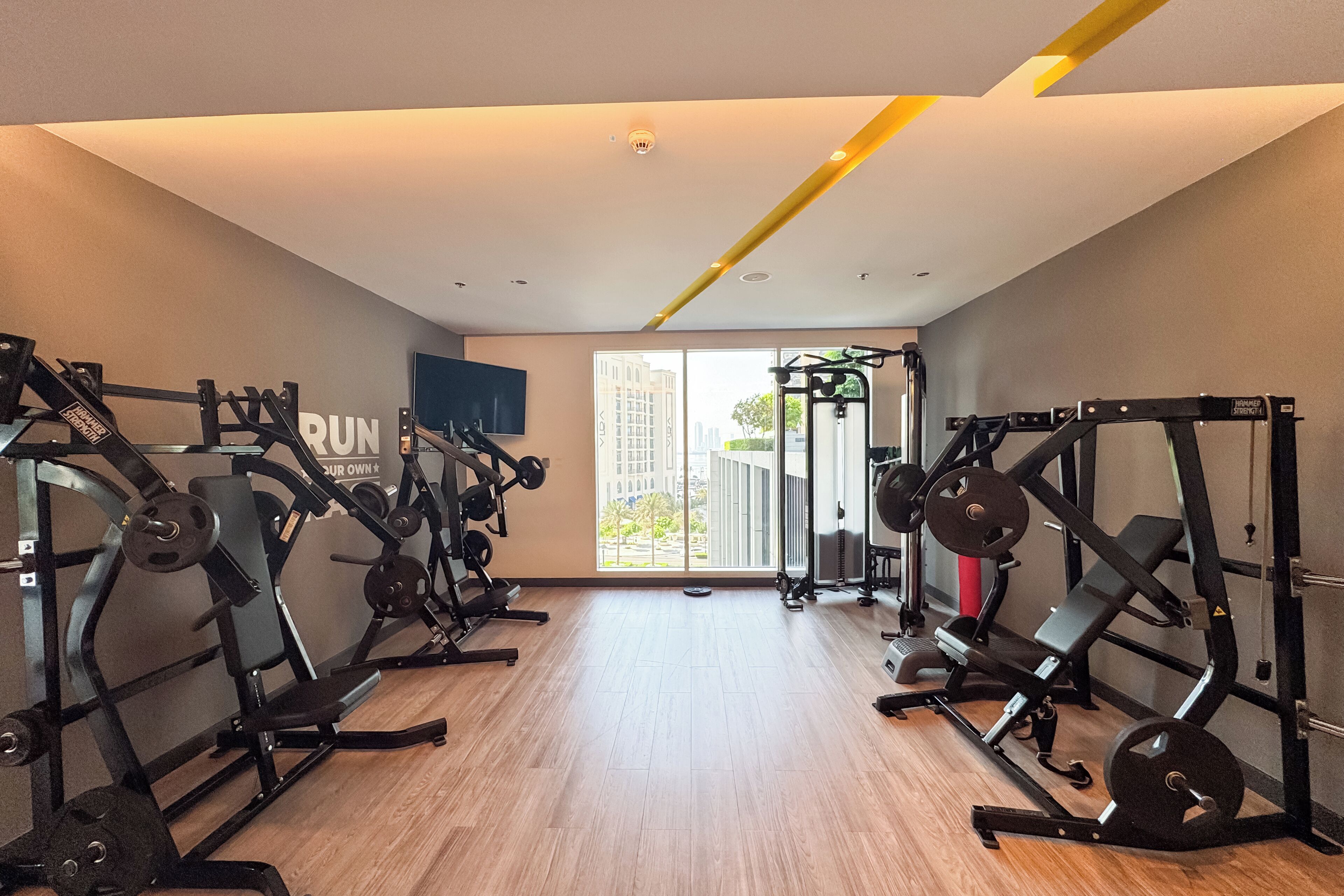 A gym with various weightlifting equipment, a large window with a view of buildings, and a flat-screen TV mounted on the wall.