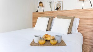 Een net opgemaakt bed met een houten hoofdeinde, twee bedlampen en een ontbijttafel met croissants en sinaasappelsap.