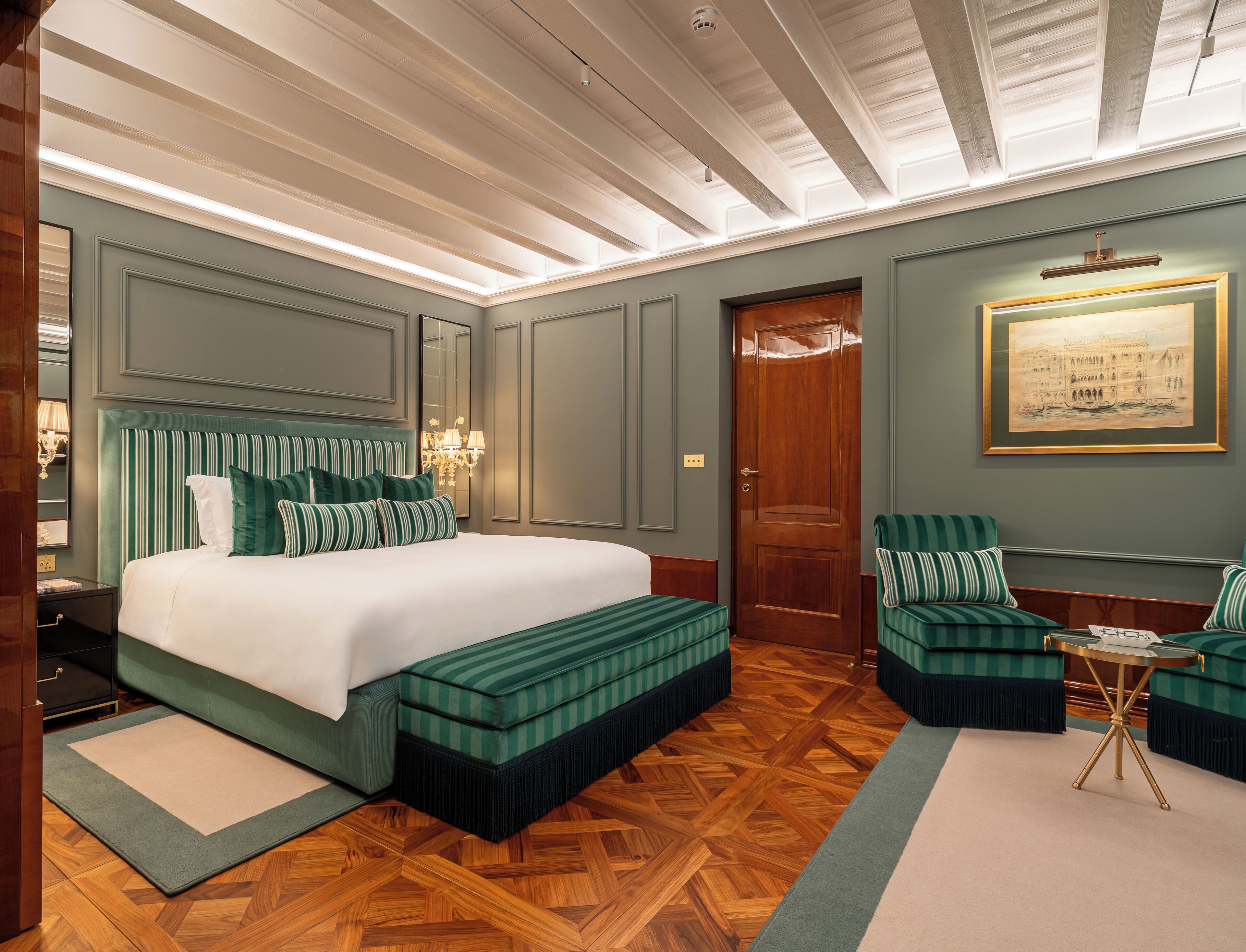 A hotel room with a large bed, a sitting area with a chair and a small table, a wooden parquet floor, and a framed picture on the wall.