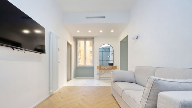 Una sala de estar moderna con un televisor de pantalla plana montado en la pared, un sofá de color claro y un suelo de madera con patrón de espiga.