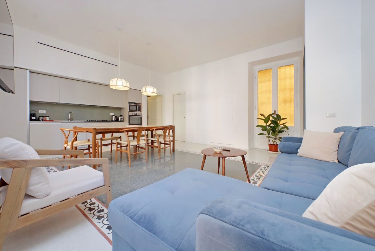 A modern living room with a blue sofa, a wooden chair, and a dining table with chairs. There is a kitchen area with cabinets and a microwave.