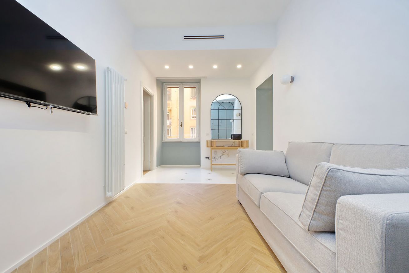 A modern living room with a flat-screen TV mounted on the wall, a light-colored sofa, and a herringbone-patterned wooden floor.