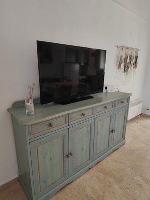A flat-screen TV on a light blue cabinet with drawers, a wall-mounted macrame decoration, and a glass vase with reeds.