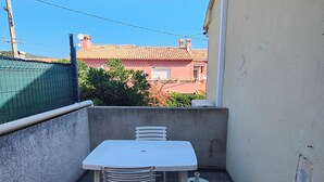A small outdoor balcony with a white table and chairs, a concrete wall, and a view of buildings in the background.