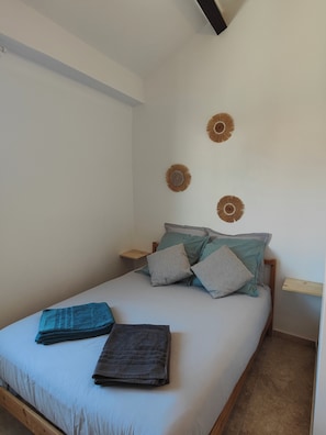 A single bed with a wooden frame, a blue and grey blanket, and two pillows. There are three wall-mounted decorative items. - A Casa Sulana—300 meters from the port of Macinaggio (Tomino)