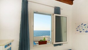 Une pièce dotée d’une grande fenêtre donnant sur la mer, d’un petit balcon avec une chaise et des plantes en pot, et d’un mur blanc orné d’une décoration sur le thème des poissons.