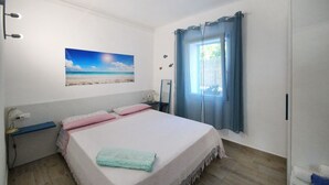 Een slaapkamer met een bed, nachtkastje, lamp, raam met gordijnen en een schilderij van een strand.