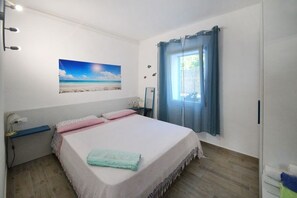 A bedroom with a bed, a nightstand, a lamp, a window with curtains, and a beach scene painting on the wall. - The Casello sul Mare is a historic railway house built in 1895 as a residence for railway workers and restored in 2025 while preserving its authentic charm. The property is set in a truly unique location, nestled between the railway and the sea, offering (Torremuzza)