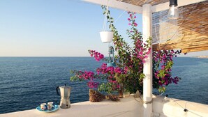 Une terrasse en bord de mer offrant une vue sur la mer, un pot suspendu orné de fleurs roses, une cafetière et une table avec une théière et des tasses.