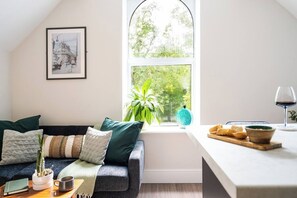 A cozy living room with a grey sofa, a window with a view of greenery, a framed picture on the wall, and a table with a wine glass and bread. - 2-Bed · City Centre · Sleeps 4 (Cardiff)