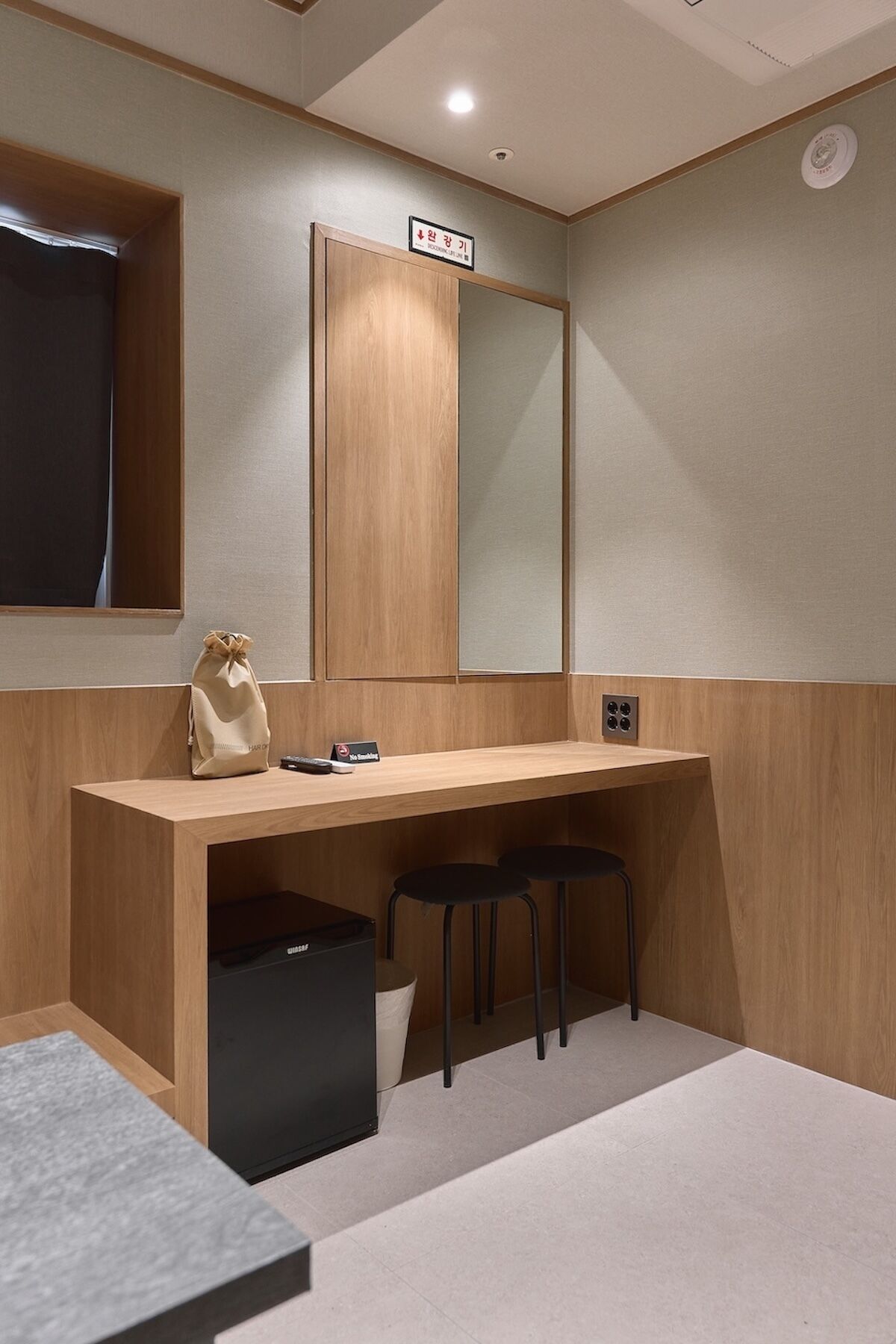 A modern bathroom with a wooden vanity, a mirror, a small trash bin, and two stools.