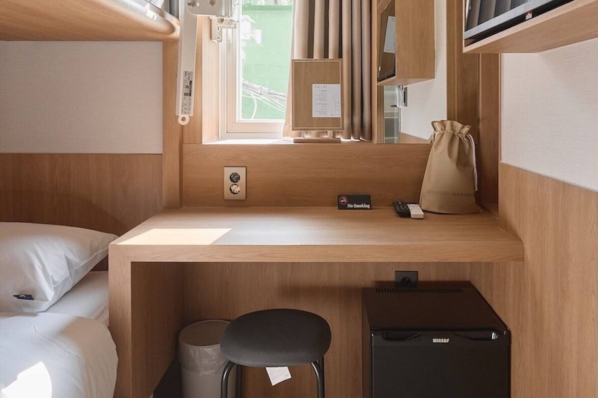 Een kleine kamer met een stapelbed, een klein bureau met een stoel, een raam met gordijnen en een compacte koelkast.