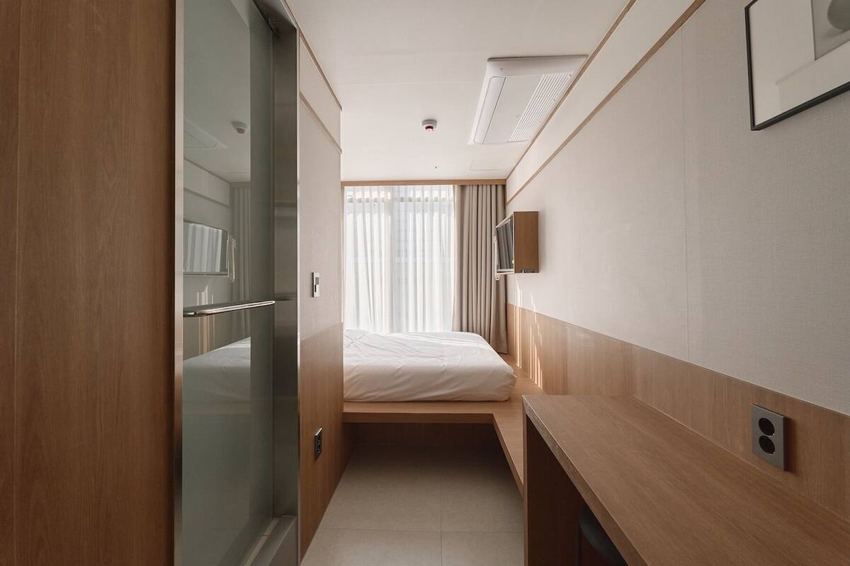 Een smalle hotelkamer met een bed, een klein bureau, een spiegel en een televisie.