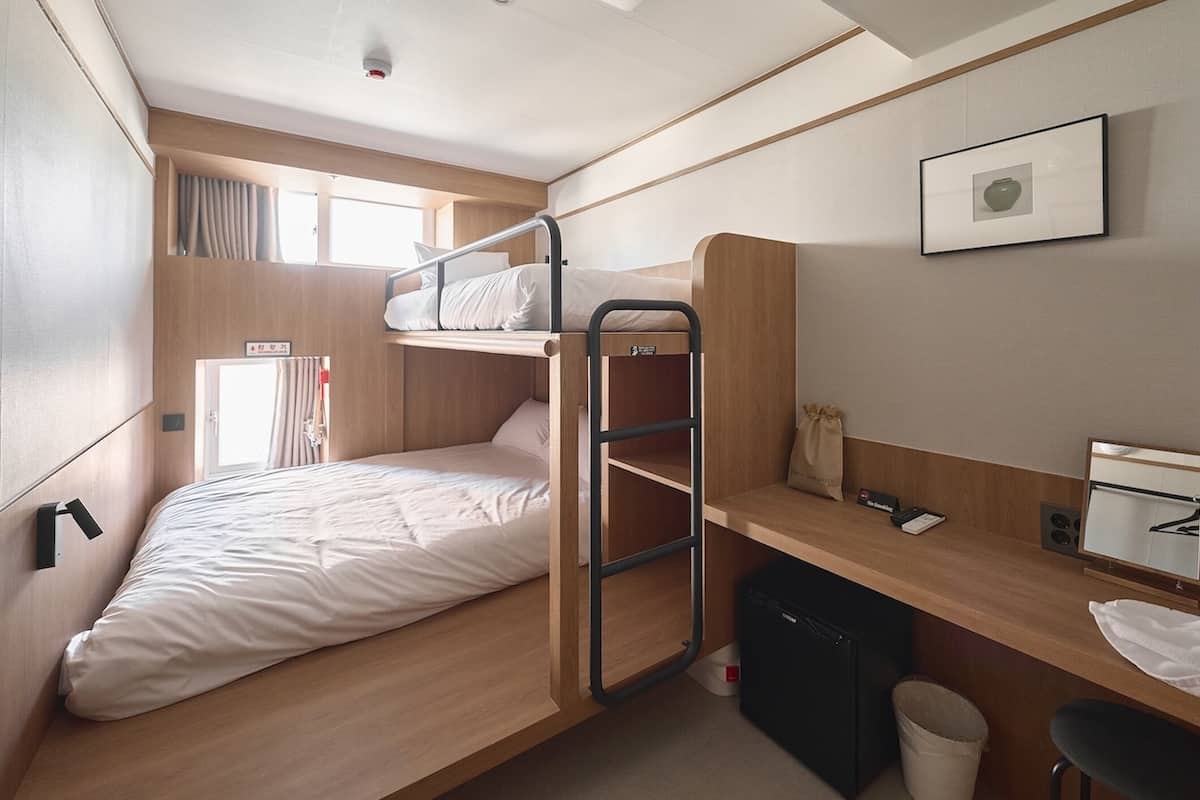 Een compacte hotelkamer met een stapelbed, een bureau met stoel, een kleine prullenbak en een raam met gordijnen.