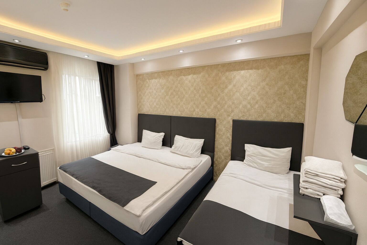 A modern hotel room with a large bed, a bedside table with a lamp, a flat-screen TV mounted on the wall, and a small table with fruits and a phone.