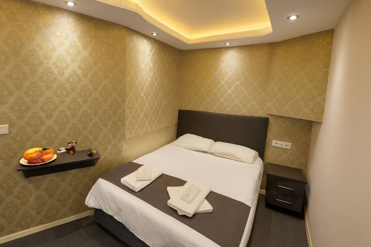 Habitación de hotel con cama, mesitas de noche, una estantería con frutas y una pared con diseño decorativo.