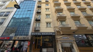 Un hotel llamado HOTEL SEZGIN con fachada de vidrio moderna, un edificio tradicional con balcones y una tienda con un letrero que dice "NAPLE COFFEE".