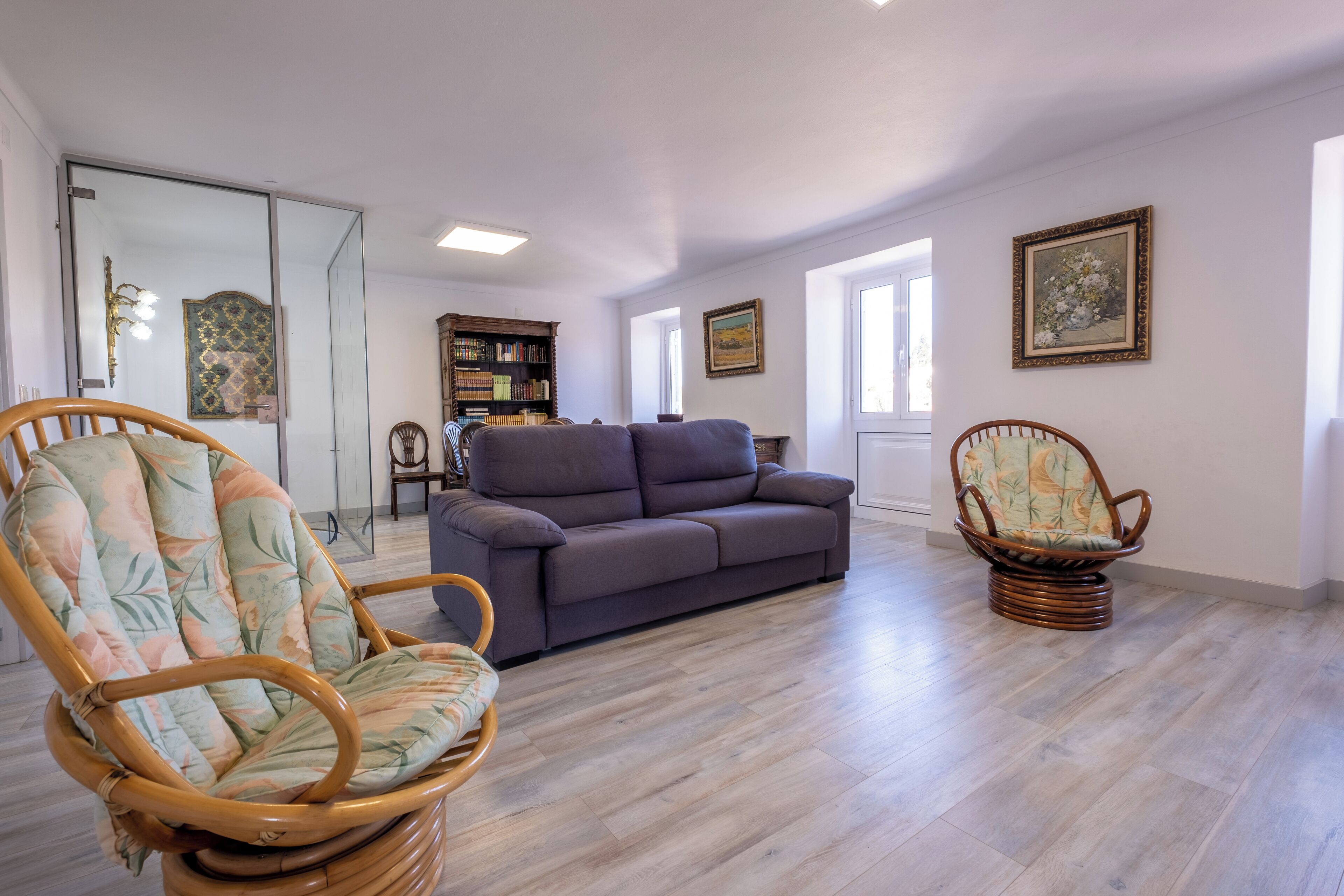 Un salón espacioso con un sofá gris, dos sillones de mimbre, una estantería y un espejo grande.