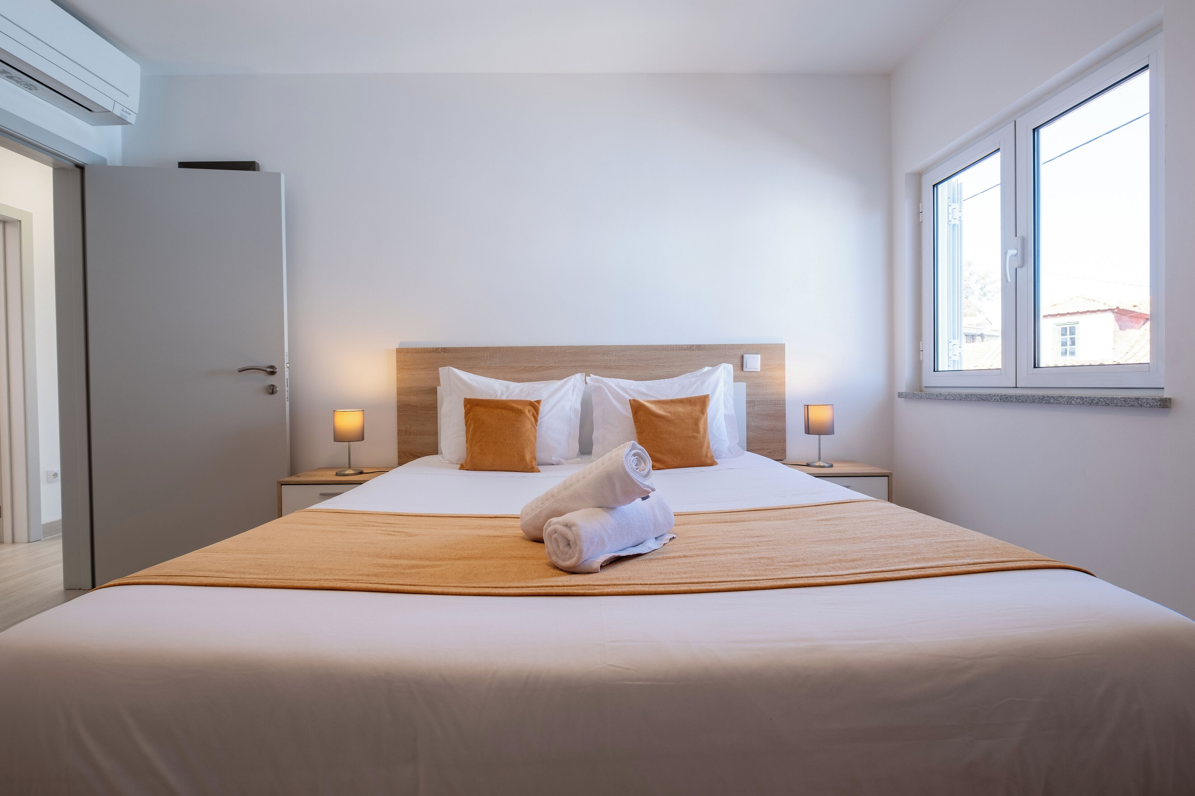 Una cama bien hecha con cabecera de madera, dos lámparas de noche y una ventana con vista a edificios.