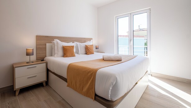 A modern bedroom with a large bed, a nightstand with drawers, a bedside lamp, and a view of the sea through a large window.
