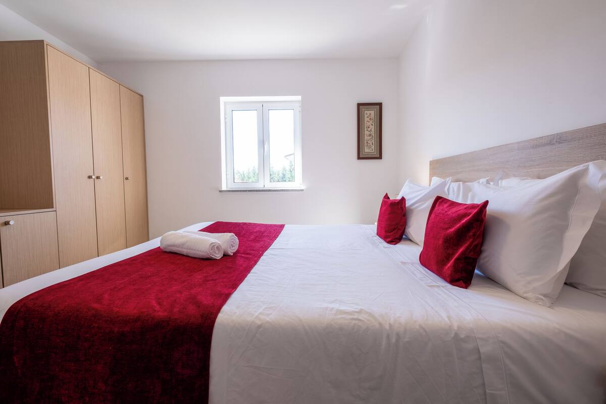 A neatly made bed with a red blanket and pillows, a window with blinds, a wooden wardrobe, and a framed picture on the wall.