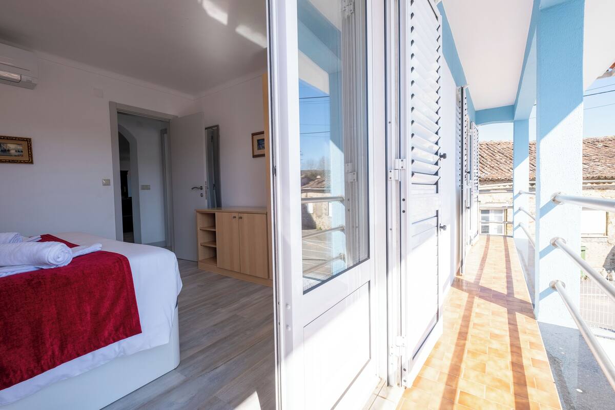 A hotel room with a bed, a red blanket, a wooden wardrobe, and a balcony with a railing.
