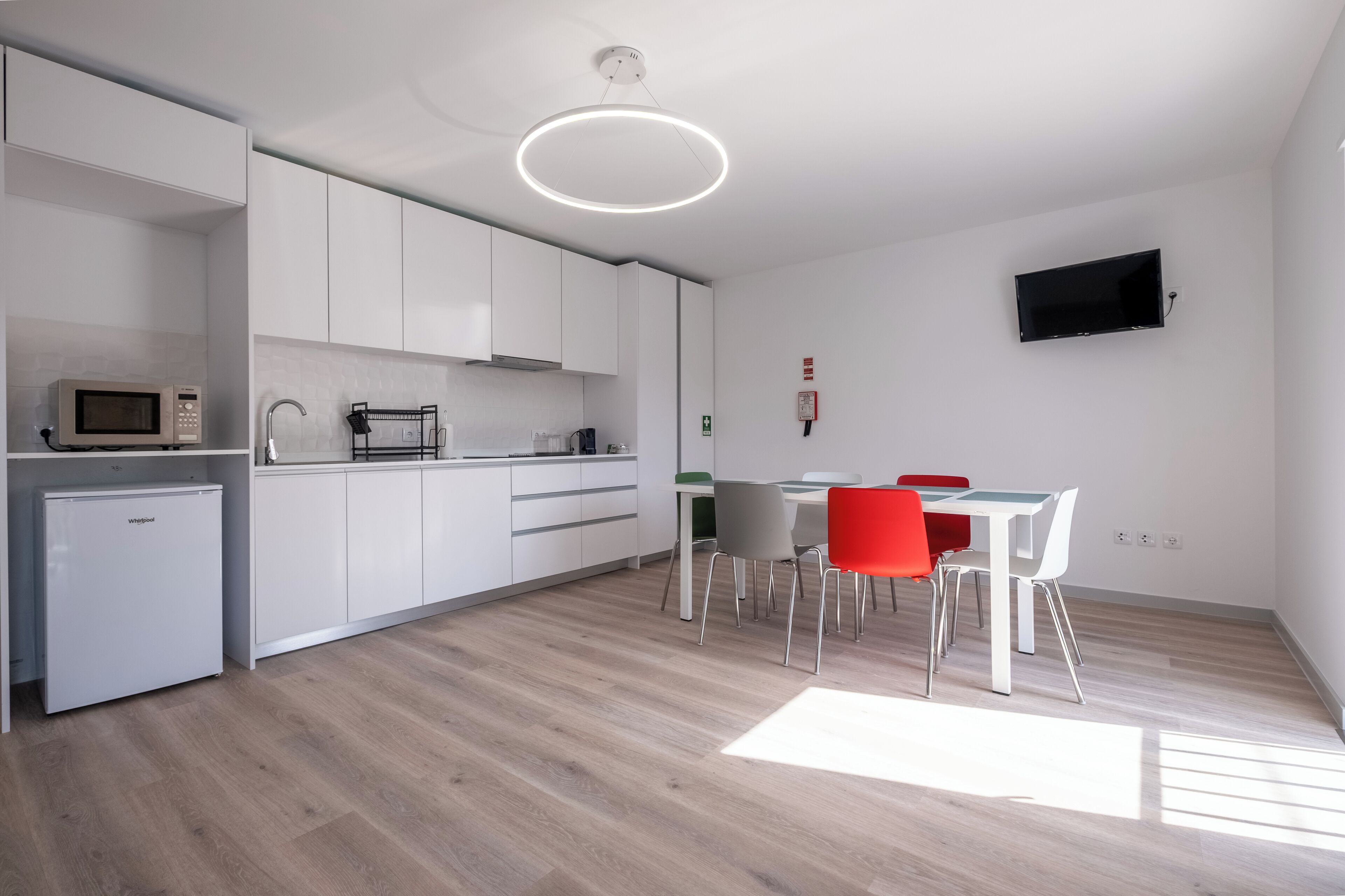 A modern kitchen with white cabinets, a microwave, and a small refrigerator. There is a dining area with a table and chairs, and a flat-screen TV mounted on the wall.