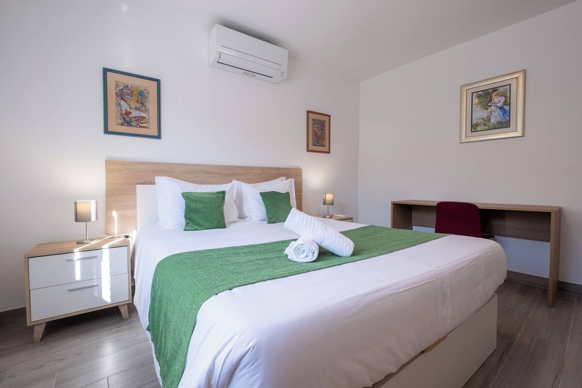A neatly made bed with a green blanket, a wooden headboard, and a white pillow. There is a desk with a red chair and a lamp.