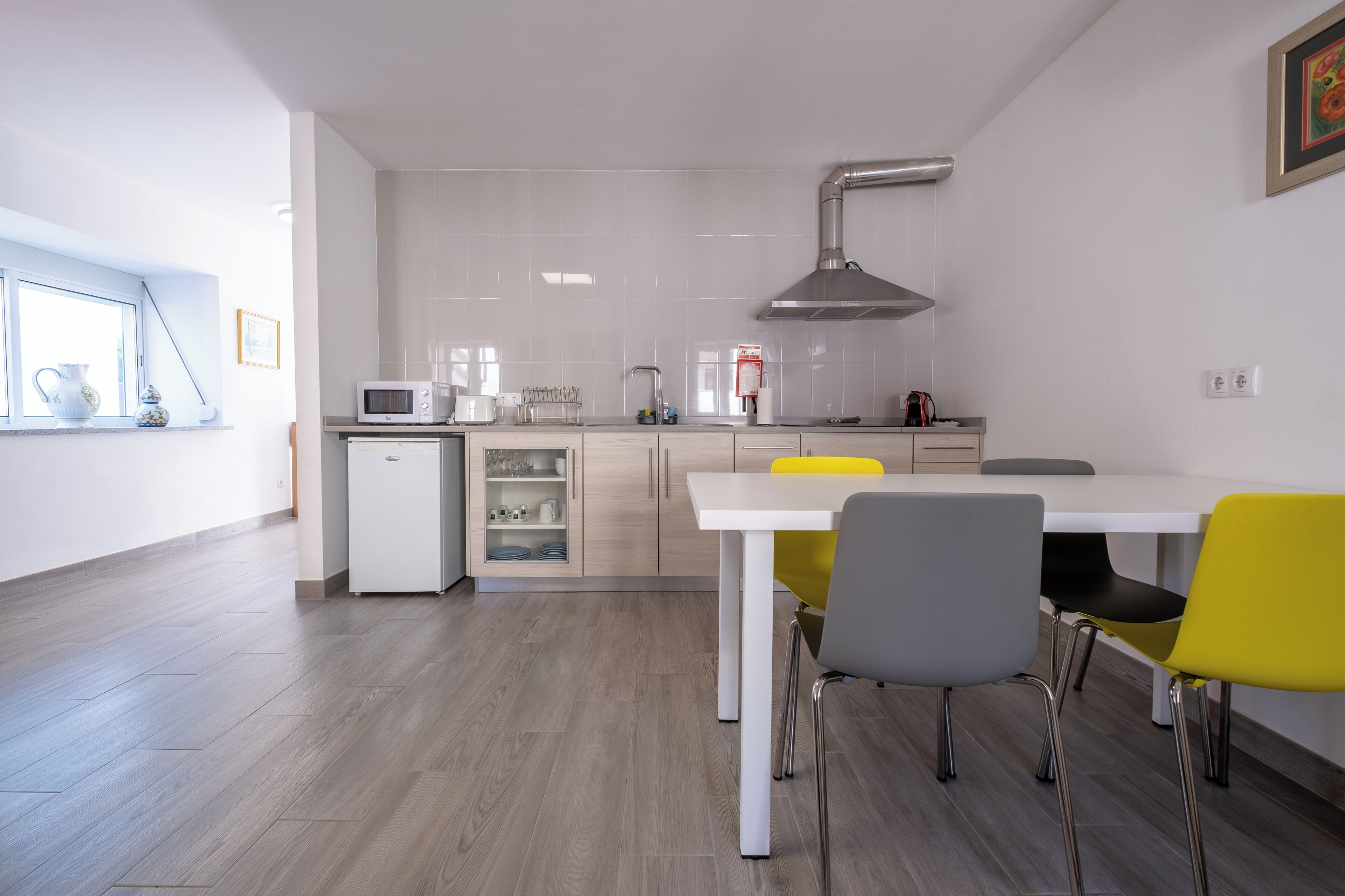 Une cuisine moderne avec un plan de travail blanc, un four à micro-ondes et un évier. On y trouve une table à manger avec des chaises et une fenêtre qui laisse entrer la lumière naturelle.
