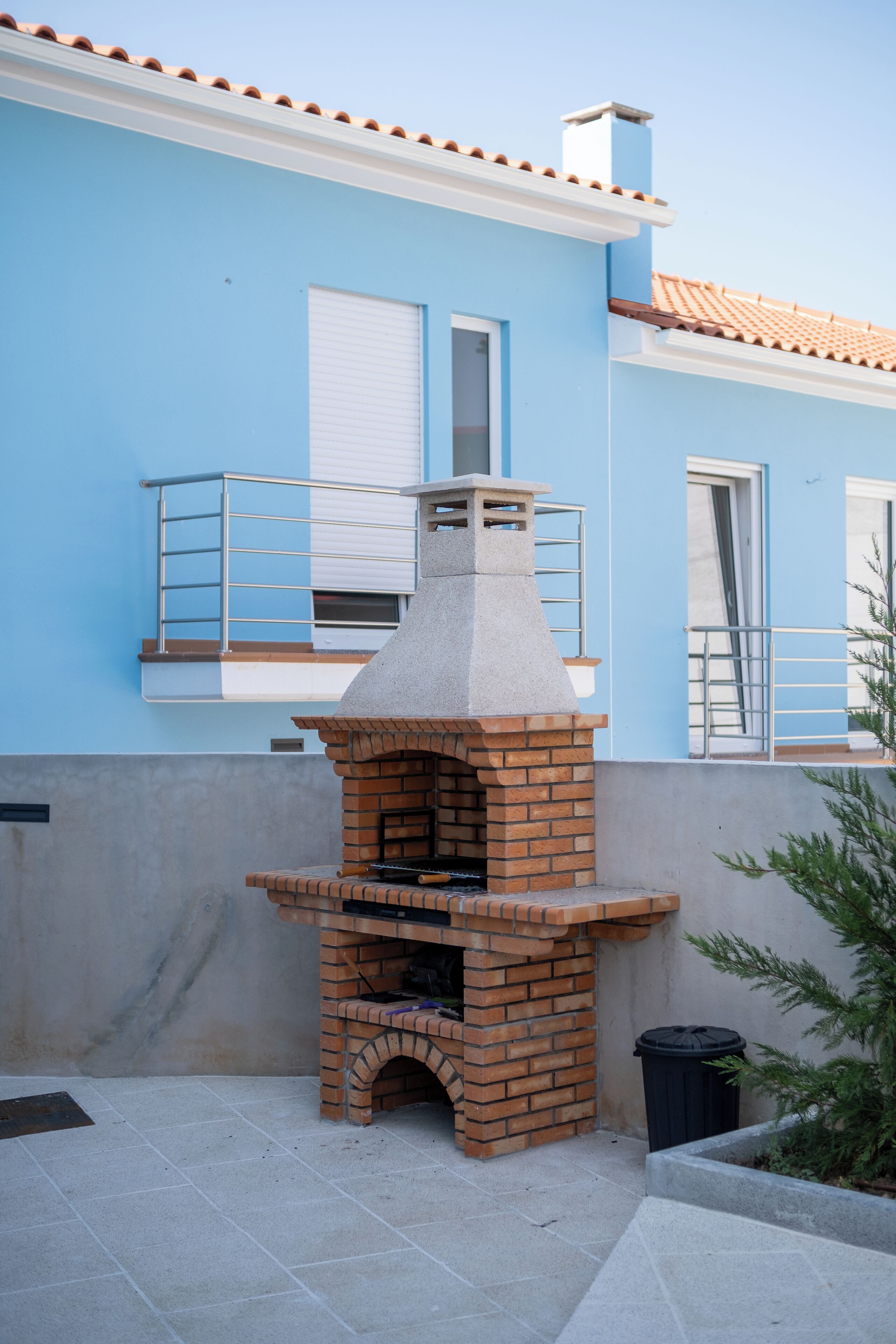 A brick outdoor oven against a blue building with a tiled roof and a window with a white shutter.