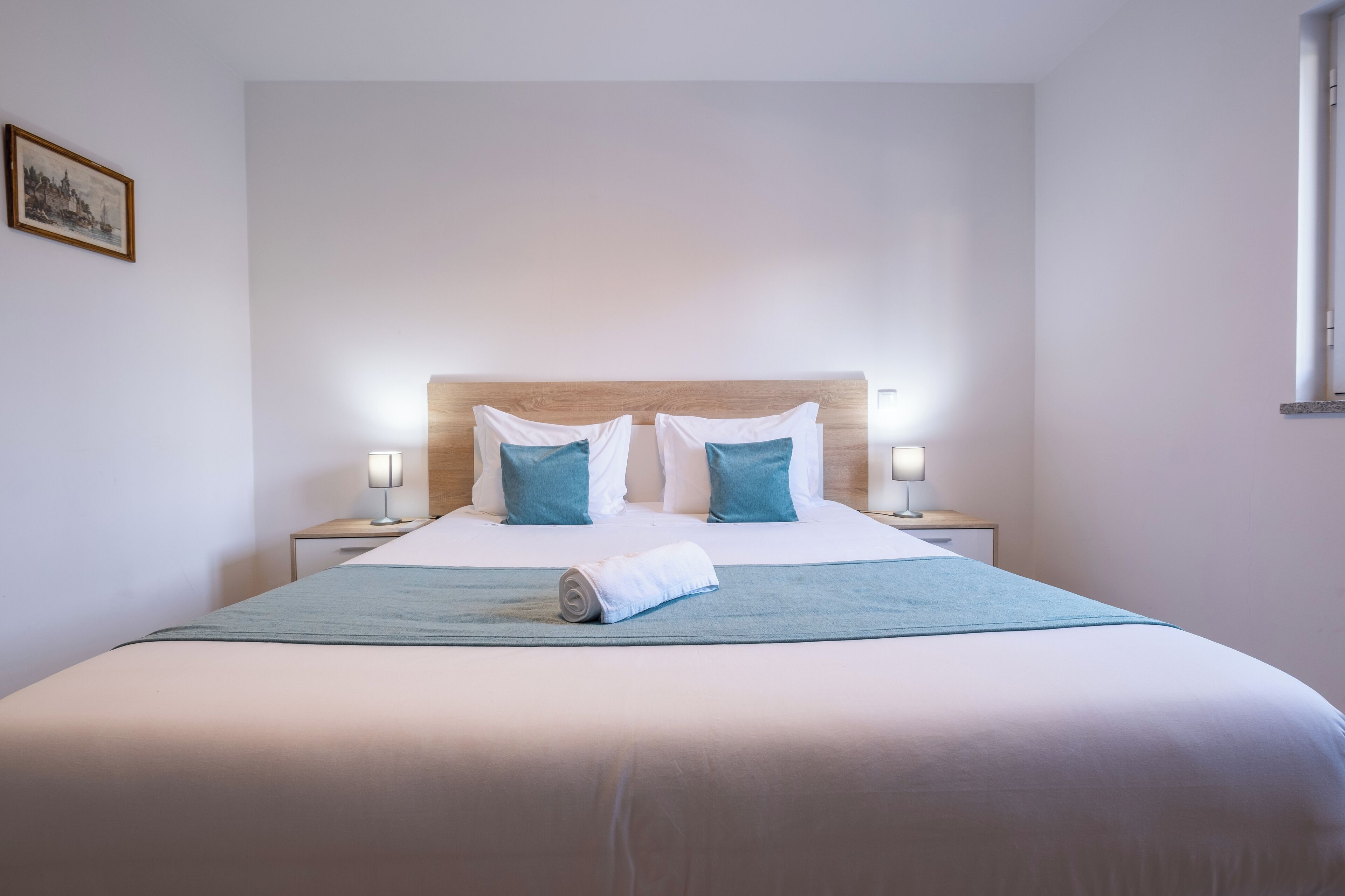 A neatly made bed with a wooden headboard, two bedside lamps, and a rolled towel on the bed.