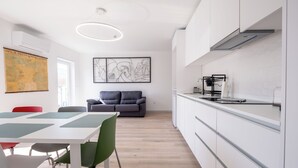 A modern kitchen with white cabinets, a dining area with a white table and green chairs, and a gray sofa in the background.