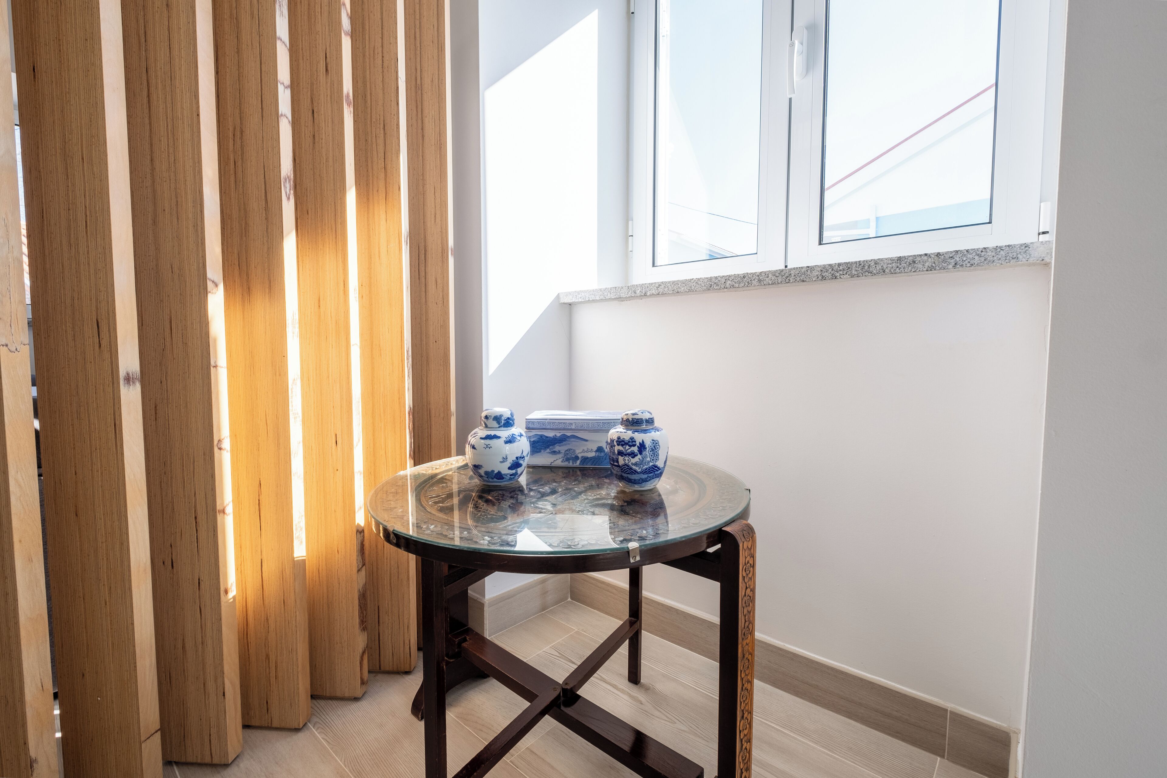 Une table ronde en verre avec un cadre en métal foncé, deux vases en céramique bleue et blanche, et un paravent en lattes de bois.