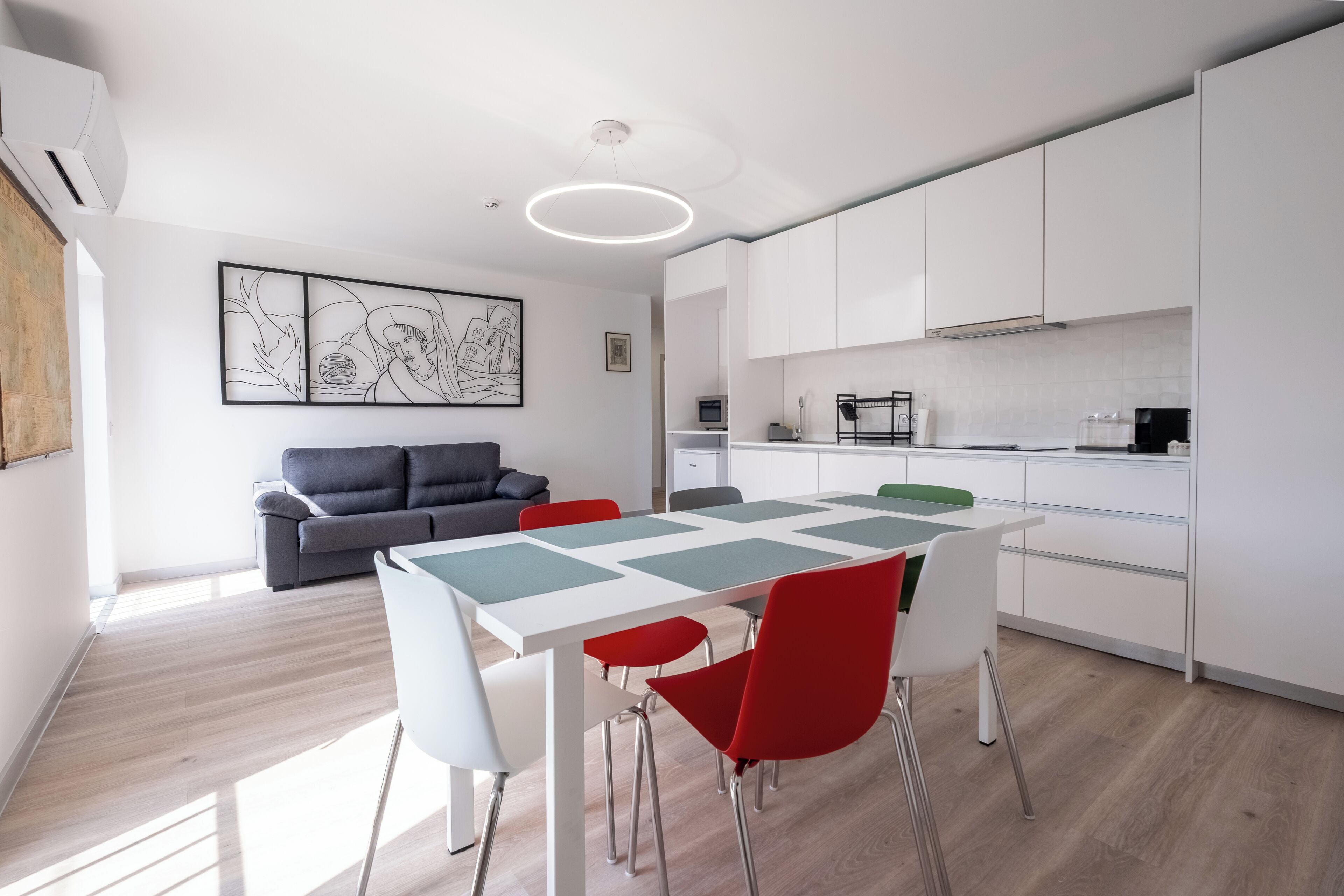 A modern kitchen with white cabinets, a dining table with red chairs, and a gray sofa in a bright living area.