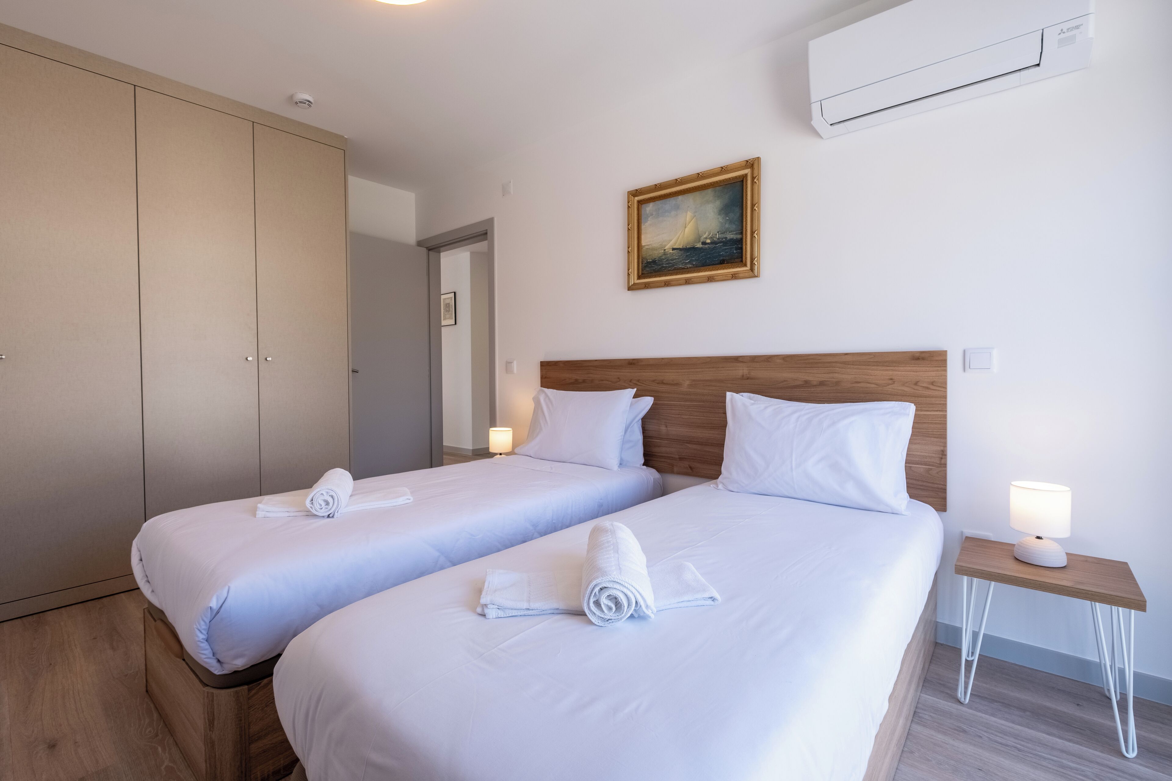 A hotel room with two beds, a wooden headboard, a nightstand with a lamp, and an air conditioning unit on the wall.