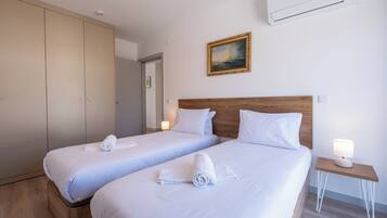 A hotel room with two beds, a wooden headboard, a nightstand with a lamp, and an air conditioning unit on the wall.