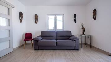 En moderne stue med en grå sofa, en rød lænestol og et lille bord med en lampe. Fire træmasker pryder væggene, og et vindue er dækket af skodder.