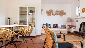 Ein Wohnzimmer mit Rattanmöbeln, einer Couch mit bunten Kissen, einer Essecke mit Tisch und Stühlen sowie einem dekorativen Wandbild.