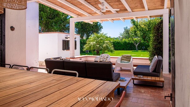 Eine Terrasse im Freien mit einem Holztisch, einer Couch und einem Sessel. Ein Deckenventilator und ein Blick auf einen Garten sind vorhanden.