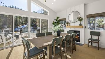 A spacious dining area with a large wooden table and chairs, a fireplace, and large windows offering a view of the outdoors.
