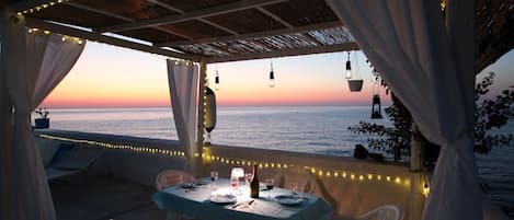 An outdoor dining area with a table set for two, overlooking the sea at sunset.