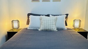 Una cama con una colcha gris oscuro, dos almohadas blancas y un cabecero negro. Hay dos mesitas de noche con lámparas de cable. Tres cuadros enmarcados están colgados en la pared sobre la cama.