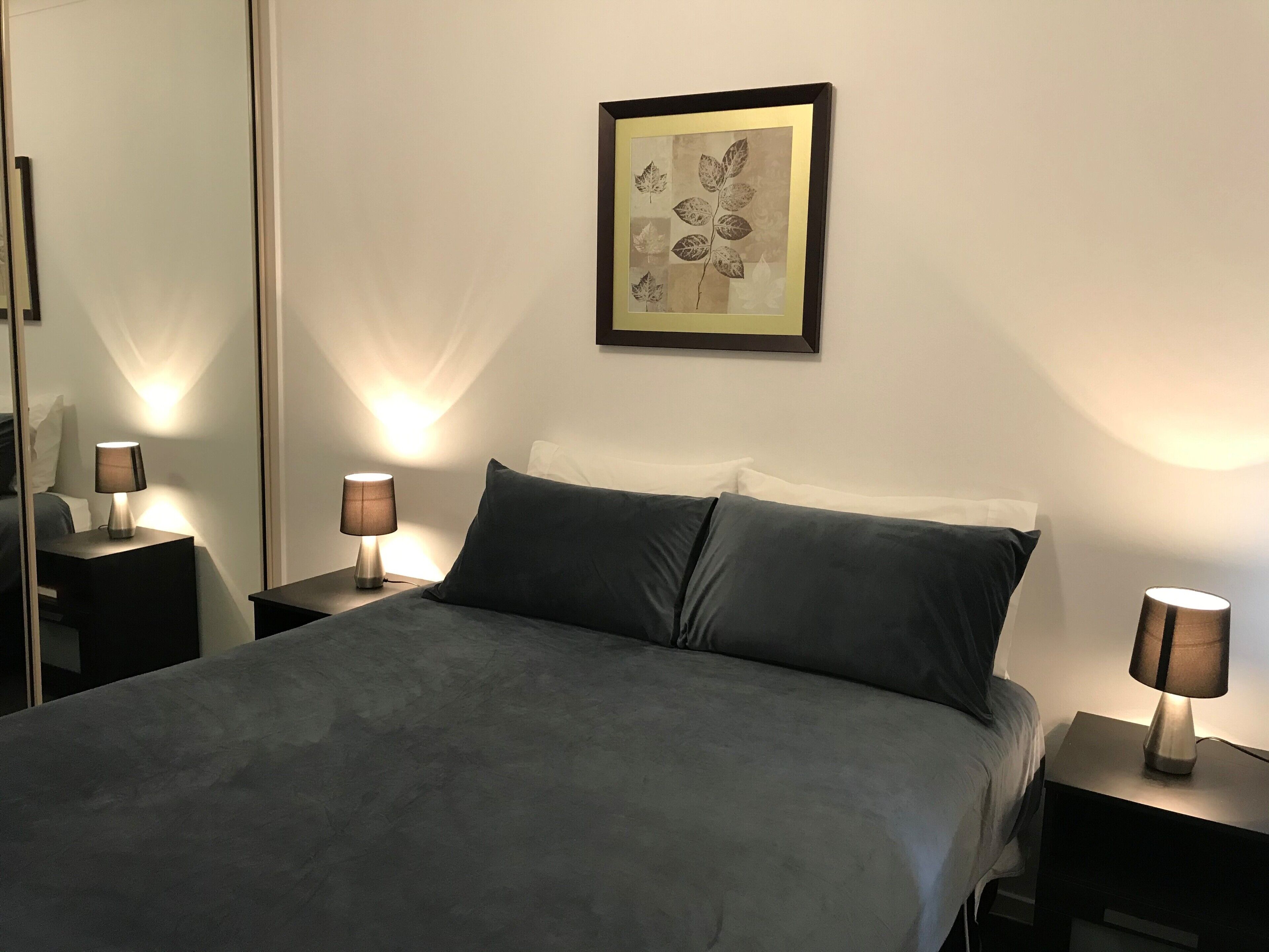 Un letto rifatto con un copriletto grigio, due comodini con lampade, un quadro appeso al muro e uno specchio a sinistra.
