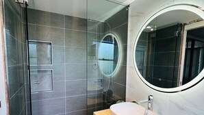 Un baño moderno con cabina de ducha de vidrio, un espejo redondo y un lavabo color beige.