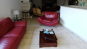 Ein Wohnzimmer mit einem roten Sofa, einem roten Sessel, einem Couchtisch, einer Chandelierleuchte und einem Küchenbereich im Hintergrund.