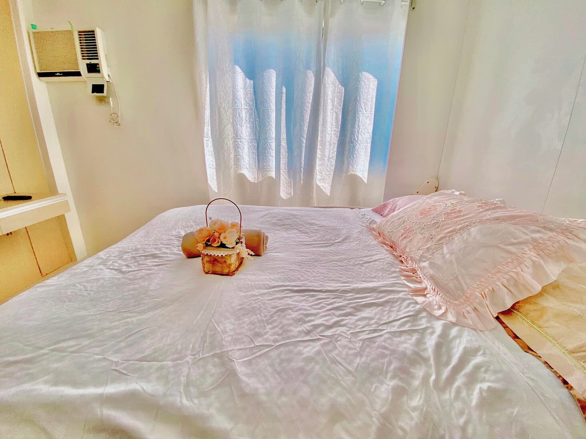 A neatly made bed with a decorative lantern, white curtains, and a wall-mounted air conditioner.